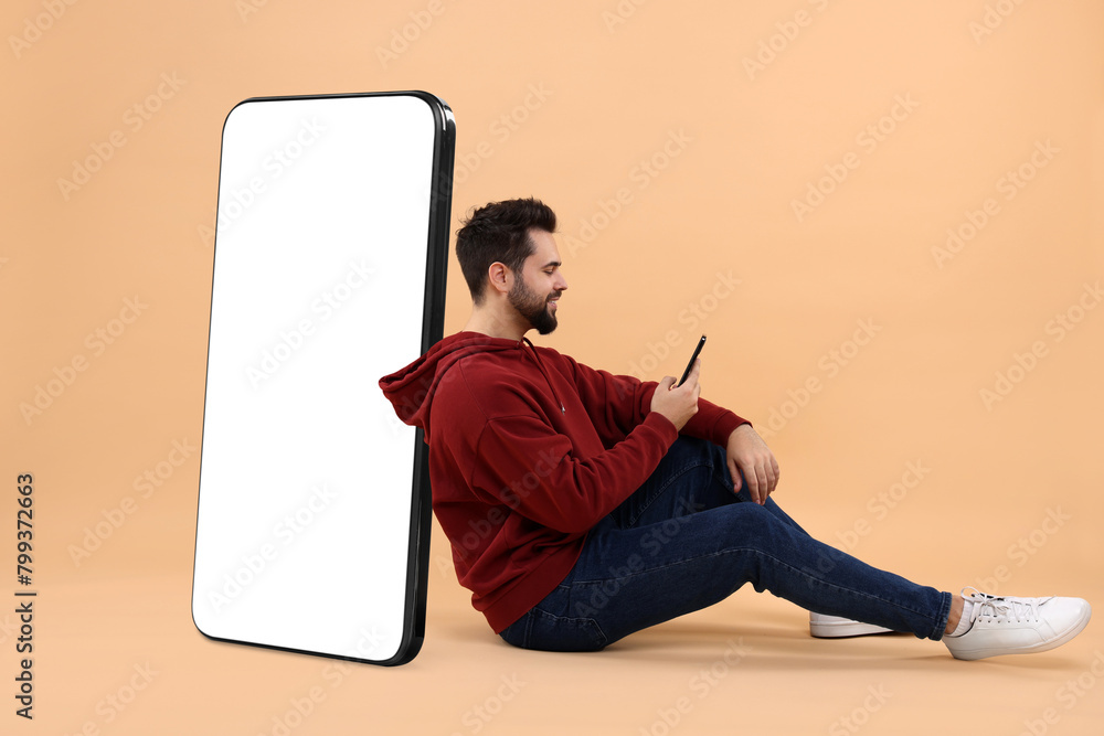 Poster man with mobile phone sitting near huge device with empty screen on dark beige background. mockup fo