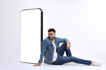 Man sitting near huge mobile phone with empty screen on white background. Mockup for design