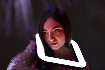 Fashionable portrait of beautiful woman wearing transparent coat with square lamp on dark background in neon lights