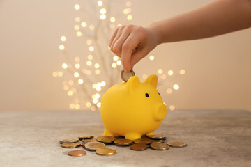 Child putting coin into piggy bank at grey table against blurred lights, closeup