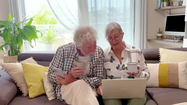 Relaxed senior couple sitting on home sofa holding a coffee cup using laptop enjoying technology, elderly 70 years old caucasian people sitting in living room browsing, shopping on line, following soc