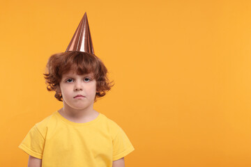 Upset little boy in party hat on orange background. Space for text