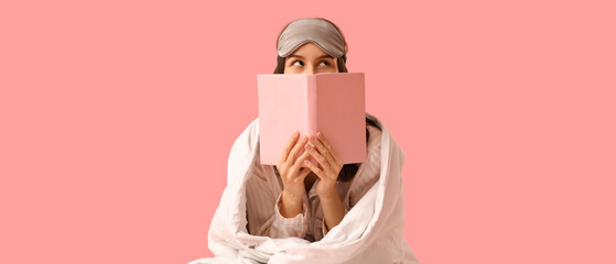 Pretty young woman with blanket, sleeping mask and reading book on pink background