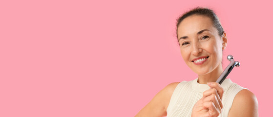 Beautiful mature woman using face massage roller on pink background