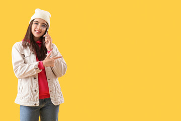 Pretty young woman talking by phone and pointing at something on yellow background