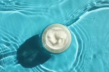 Jar with moisturizing cream in water on light blue background, top view