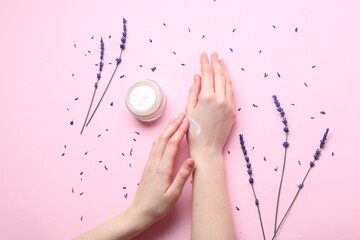 Woman applying hand cream and lavender flowers on pink background, top view