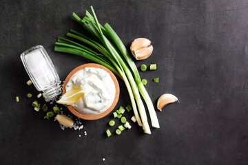 Delicious yogurt in bowl, green onion, garlic and salt on black background, flat lay. Space for text