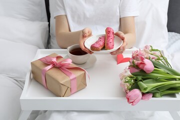 Tasty breakfast served in bed. Woman with eclairs, tea, gift box and flowers at home, closeup