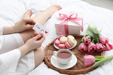 Tasty breakfast served in bed. Woman with I Love You card, macarons, tea, flowers and gift box at home, closeup