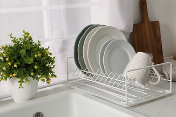 Drainer with different clean dishware, cup and houseplant on light table indoors