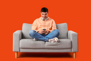 Handsome young man using mobile phone on grey sofa against orange background