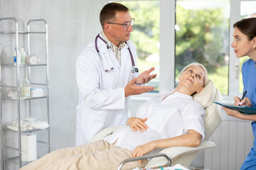 Male doctor and female assistant having conversation with elderly woman patient about need for additional diagnostic procedures, long-term and comprehensive treatment, assistant records data in report