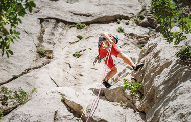 Active climber middle age man in protective helmet while abseiling from cliff rock wall using ropes with a belay device and climbing harness. Active extreme sports time spending concept