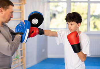 Sportive teenage boy training boxing kicks on punch mitts held by instructor in sports hall