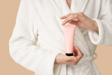 Woman in white bathrobe holding tube of cream on color background, closeup