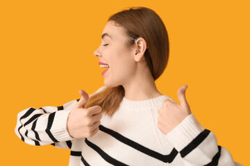 Beautiful young woman with hearing aid showing thumb-up gesture on yellow background