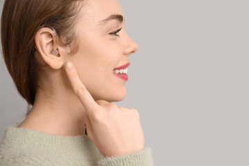 Happy young woman pointing at hearing aid on grey background