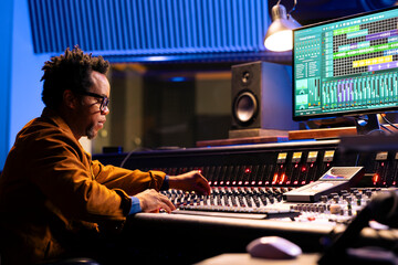 African american sound engineer works on mixing console with sliders and knobs, looking at digital...