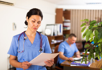 Portrait of Asian female physician in blue uniform standing with papers in medical office focused...