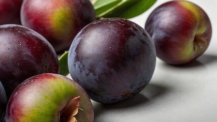 Obraz premium Several ripe plums rest on light gray surface with few green leaves scattered around them. Plums deep purple color with hints of red, green, some have visible water droplets on their skin.