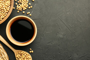 Soy sauce in bowl and soybeans on black textured table, flat lay. Space for text
