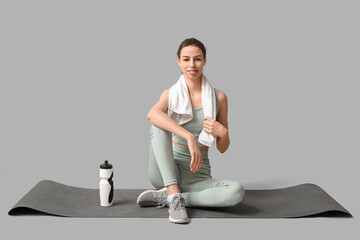 Beautiful young happy sporty woman with bottle of water and yoga mat on grey background