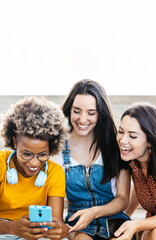 Vertical shot of three multiracial young women having fun using mobile phone sitting outdoors. Technology and social media concept.