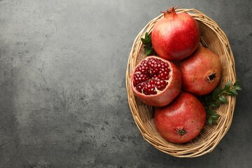 Fresh pomegranates and green leaves in wicker basket on grey table, top view. Space for text