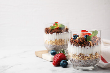 Tasty granola with berries, nuts, yogurt and chia seeds in glasses on white marble table, space for text