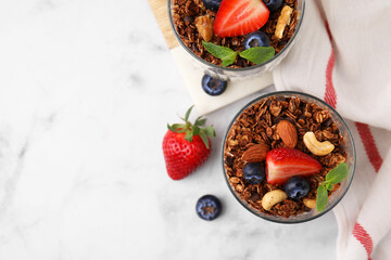 Tasty granola with berries, nuts and mint in glasses on white marble table, top view. Space for text