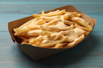Tasty potato fries and cheese sauce in paper container on light blue wooden table, closeup