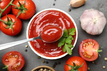 Taking delicious tomato ketchup with spoon from bowl at grey textured table with spices and products, flat lay