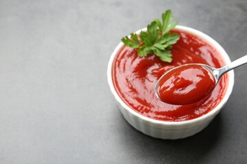 Taking delicious tomato ketchup with spoon from bowl at grey textured table, closeup. Space for text