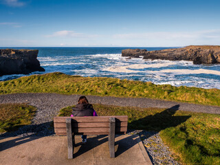 Teenager girl enjoy stunning nature of Kilkee coast and cliff, county Clare, Ireland. Rough coastline and blue cloudy sky. Travel and tourism. Irish landscape scene. Warm day. - obrazy, fototapety, plakaty