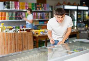 Young guy looks and chooses frozen food in refrigerator in supermarket