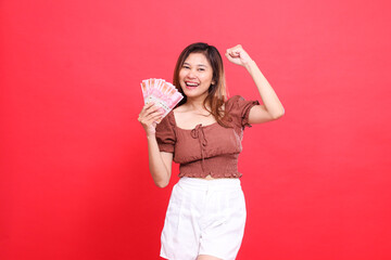 gesture of a cheerful Asian girl clenching her fist upwards successfully while holding rupiah currency wearing a brown blouse top with a red background. for transaction, technology and advertising