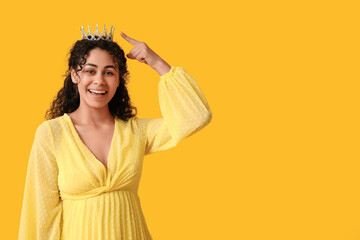 Beautiful young African-American woman in stylish prom dress pointing at tiara on yellow background