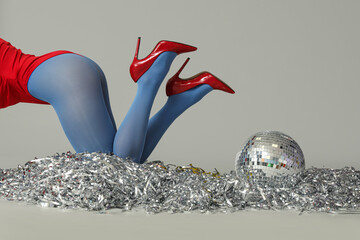 Legs of young fashionable woman with disco ball and silver tinsel on grey background, closeup