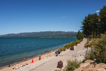 Serene Tahoe lakeside scene with sandy beach, people lounging and walking, blue lake with boats,...