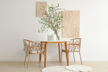 Table, armchairs and vase with blossoming branches near white wall in beautiful dining room