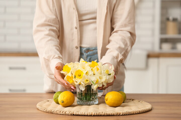 Fototapeta premium Young woman with bouquet of daffodils and fruits on table at home