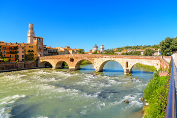 The ancient Ponte Pietra stone arch bridge crossing the Adige River, with the bell tower of the...