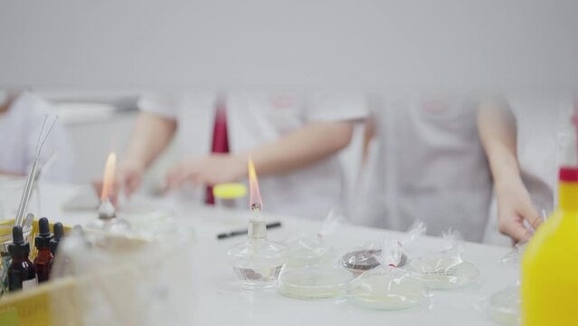 A person is holding a pair of tweezers and looking at a glass of water. The table is covered with various scientific equipment, including a beaker, a test tube, and a Bunsen burner