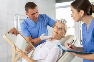 Fototapeta premium Man doctor palpates examines mature female patients facial skin and talks about need for lifting procedures to improve contour of face, female nurse records conversation data