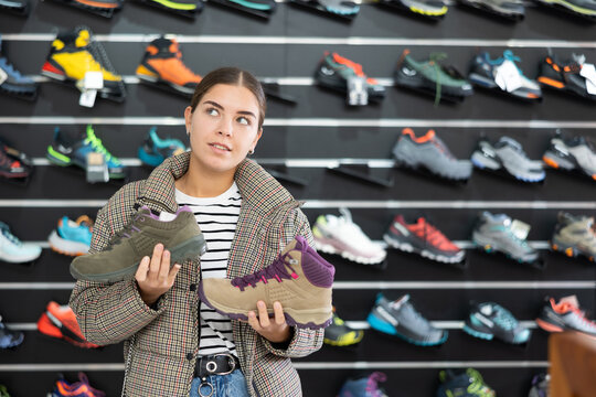 Young woman chooses specialized shoes in store, near showcase with tracking cross-shoes, buyers think about advantages and disadvantages of winter trecking boots.