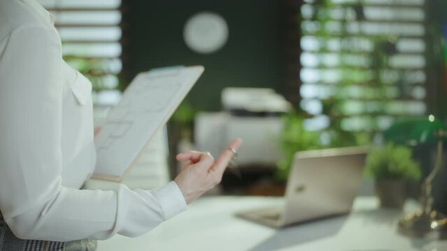 Sustainable real estate business. Closeup on young woman realtor in modern green office in white blouse with clipboard and keys.
