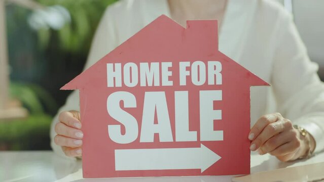 Sustainable real estate business. Closeup on middle aged woman real estate agent in green office in white blouse with home for sale sign.