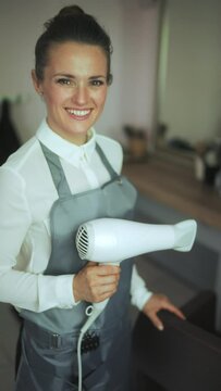 smiling middle aged woman hair salon employee in modern hair studio in uniform with hair dryer.