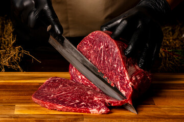 The butcher wearing black gloves cuts the meat with knife on wooden cutting board.Close-up piece of...